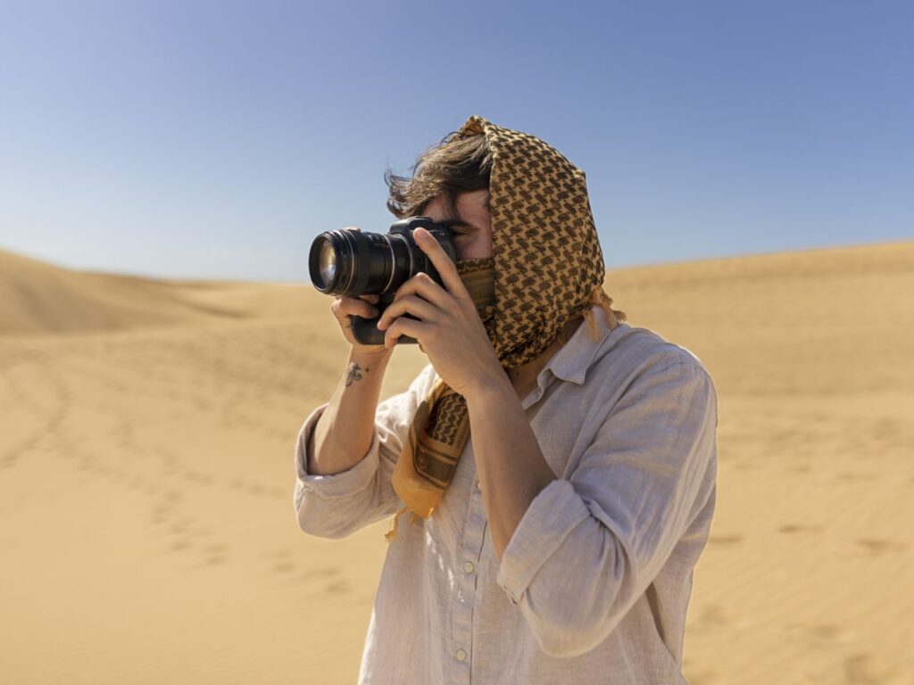 Places to visit in Egypt Cairo - Hurghada Speedboat Person wearing a patterned head scarf and light shirt, taking a photograph with a DSLR in a sandy desert landscape.